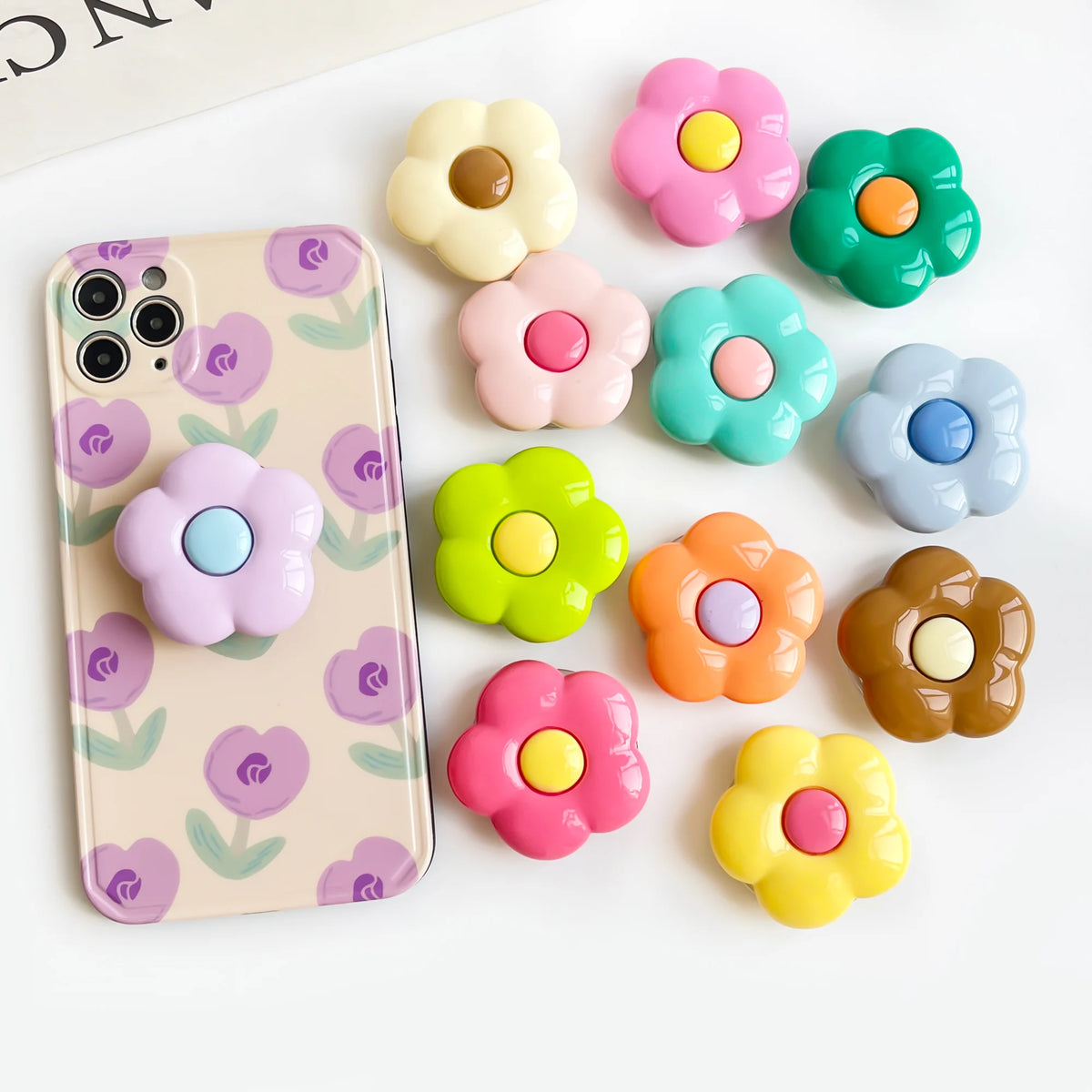 A foldable phone grip stand is shown with various colorful, flower-shaped silicone grips scattered on a white surface.