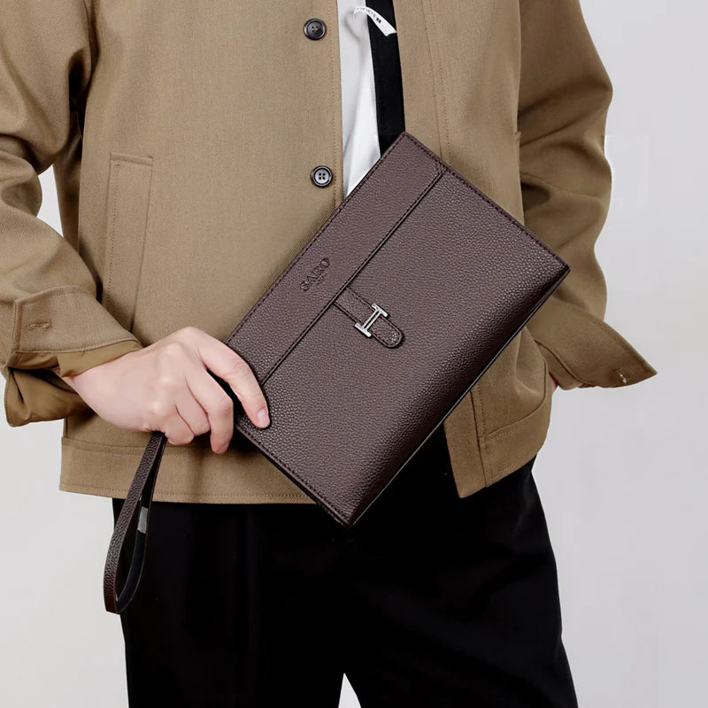 Eine Frau hält eine dunkelbraune Kunstleder-Clutch vor einem beigen Jacken und schwarzen Hosen.