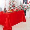 A red satin tablecloth is set for a holiday meal with festive decorations and a Christmas tree in the background.