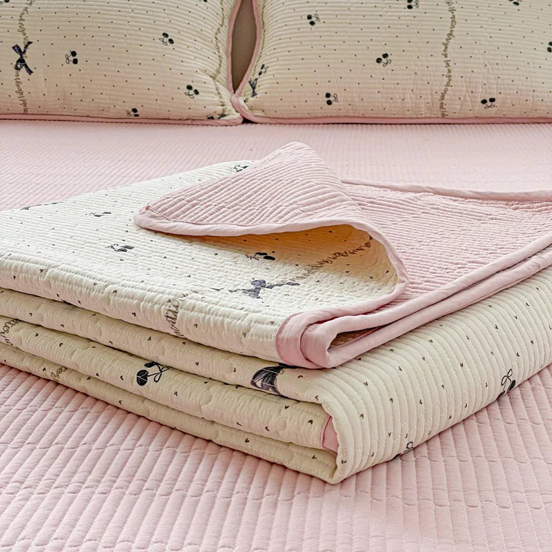 A stack of light pink and cream quilted polyester coverlets with subtle floral patterns resting on a pink surface.