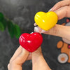 A hand holds a bright yellow heart-shaped toilet flush button and a red heart-shaped version.