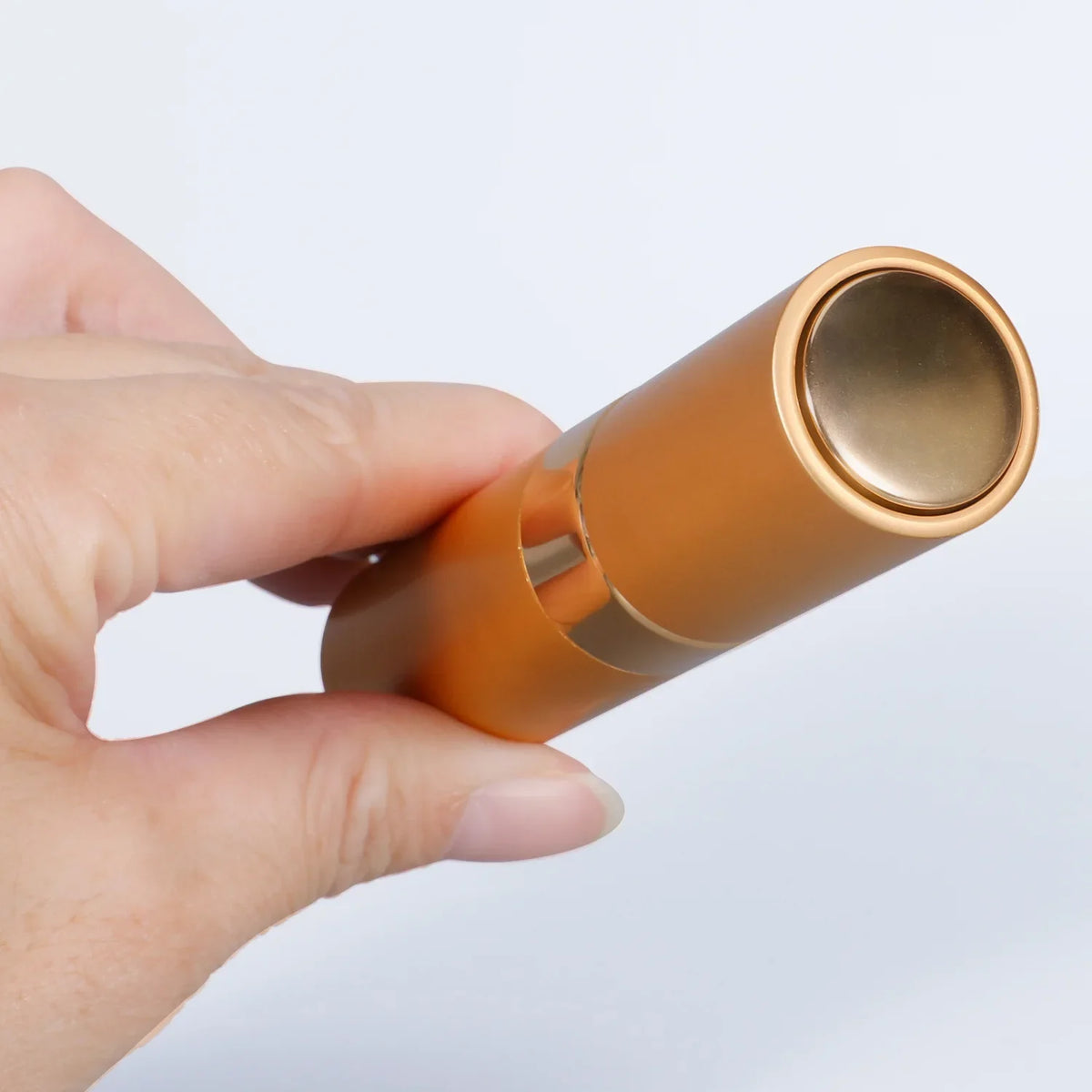 A hand holds a gold-toned refillable aluminum perfume atomizer with a silver cap.