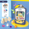A blue mini claw crane machine for kids with lights and music is displayed on a yellow stand next to various toys.