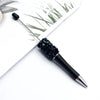 A black ballpoint pen with a sparkly diamond sequin section is shown next to some green grass and white paper.