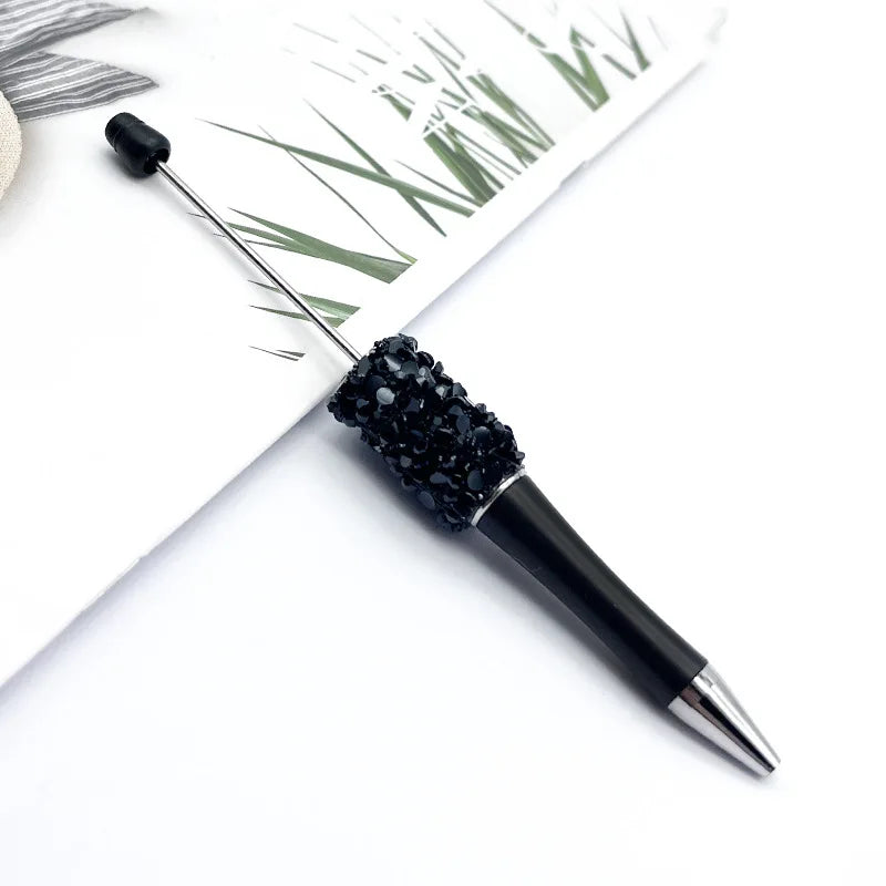 A black ballpoint pen with a sparkly diamond sequin section is shown next to some green grass and white paper.