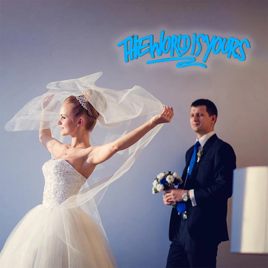 A bride in a white gown and veil is shown alongside a groom, with a neon sign reading The World Is Yours above them.