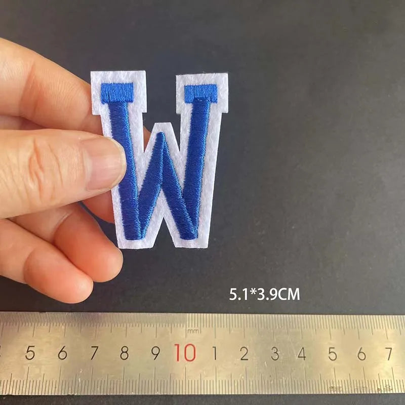 A hand holds a blue embroidered letter W patch, measuring 5.1 by 3.9 cm against a ruler.