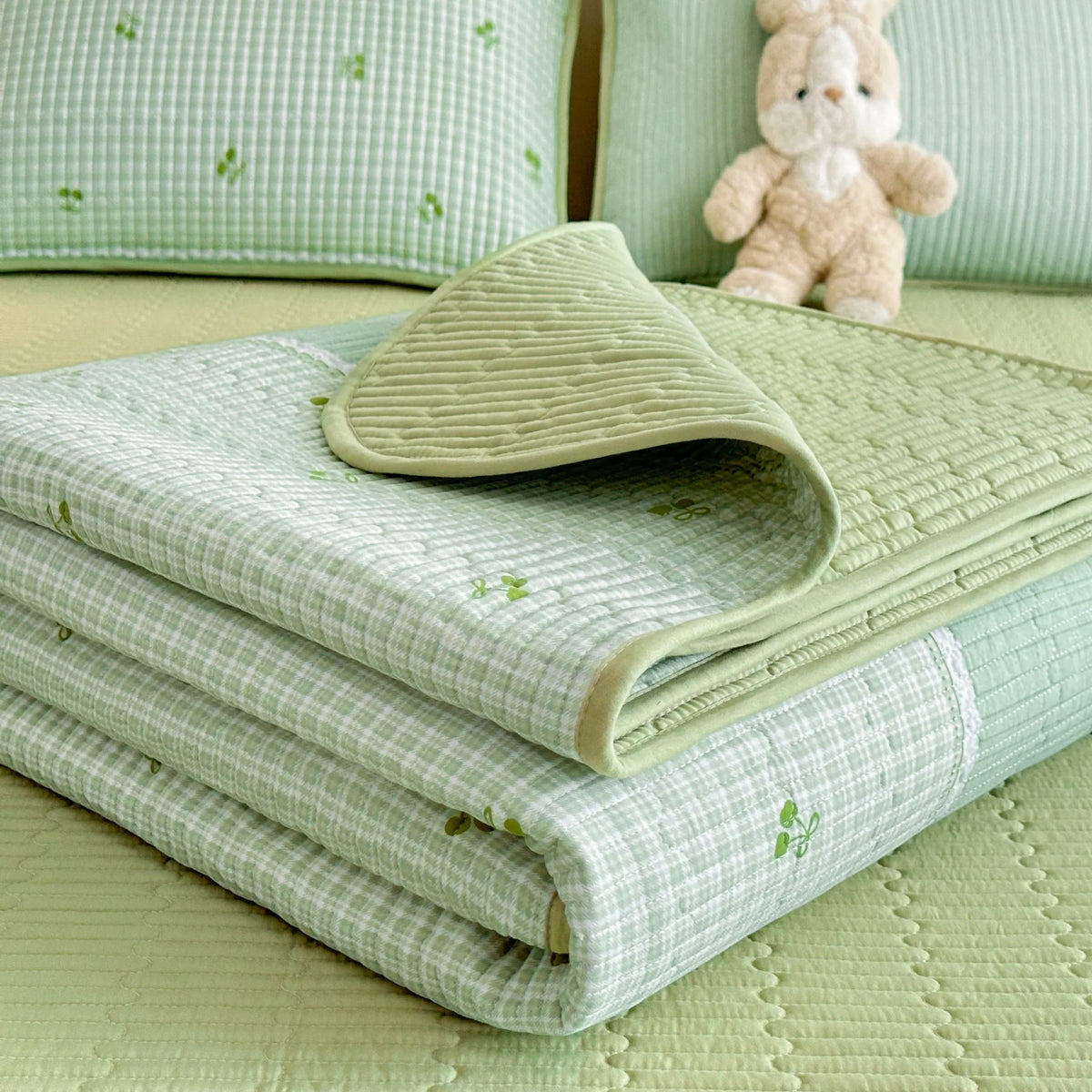A stack of light green quilted fabric and a beige dust-proof cover with a small teddy bear in the background.