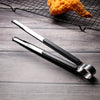 A pair of shiny stainless steel BBQ tongs resting on a dark wooden surface next to a piece of fried food on a wire rack.