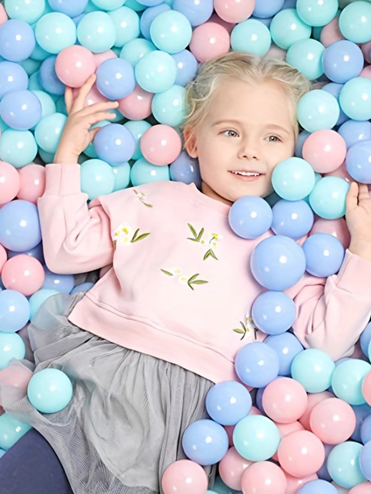 A smiling young girl is surrounded by a large collection of colorful plastic ball pit balls.