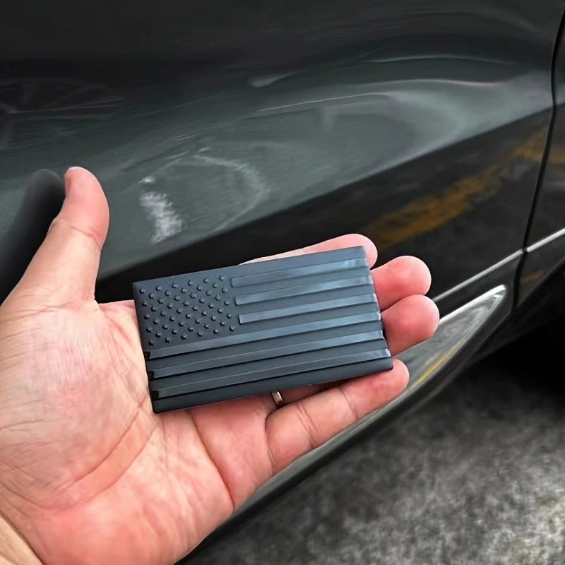A hand holds a matte black 3D American flag car emblem set with ridged detailing against a dark car surface.