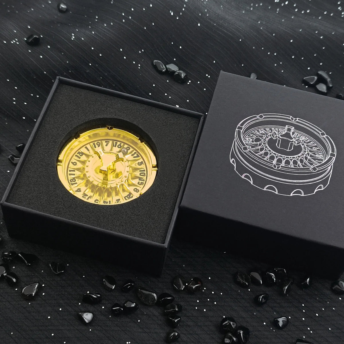 A gold metal fingertip gyro roulette dice is displayed inside a black box on a dark surface.
