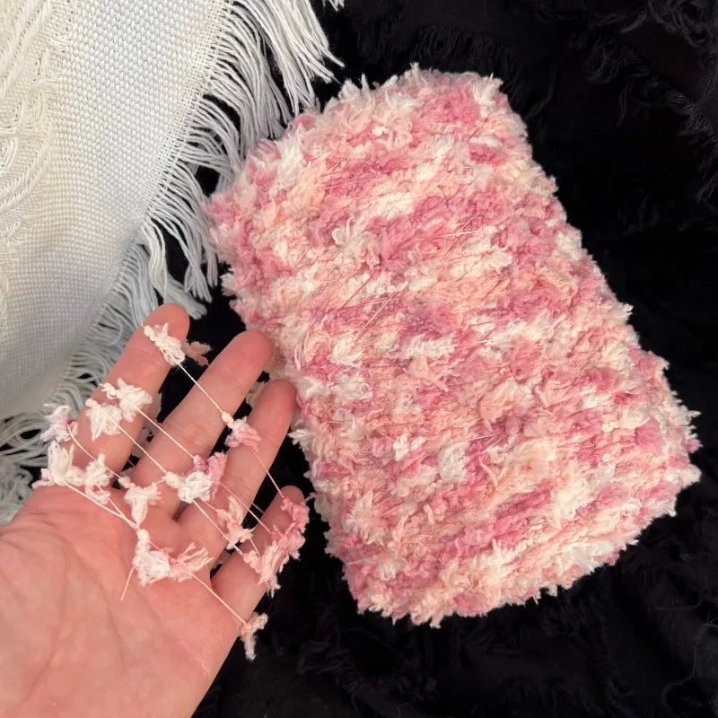 A pink variegated yarn is shown next to a hand, resting on a black fuzzy surface.