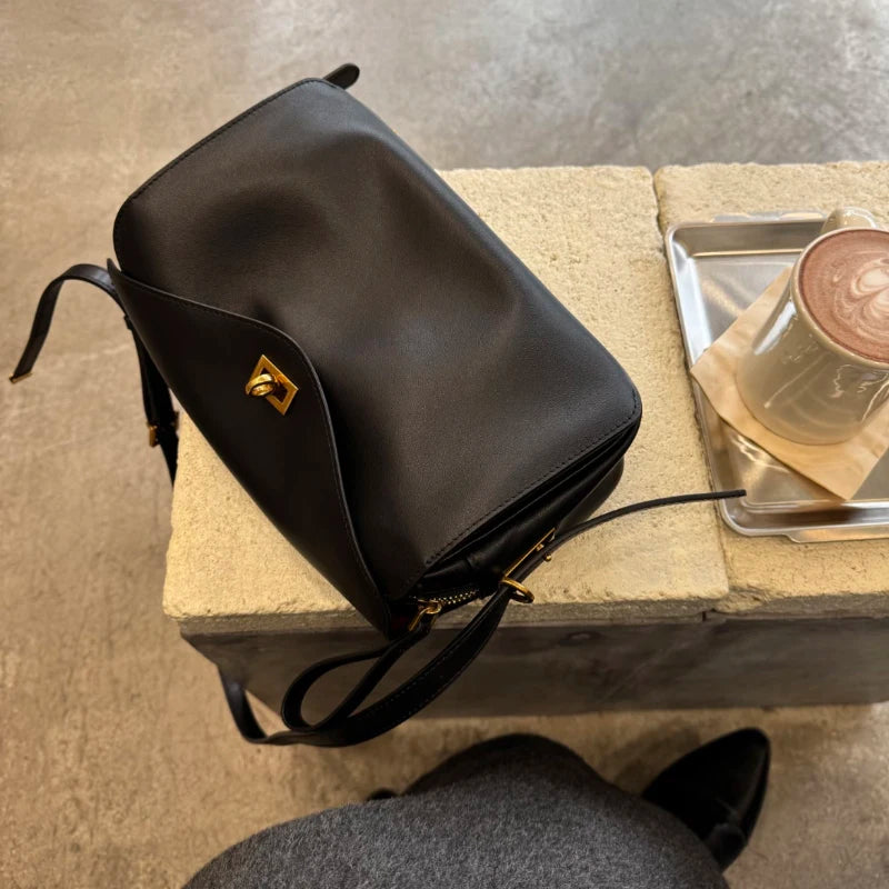 A black genuine leather square crossbody bag with a gold lock resting on a light surface.