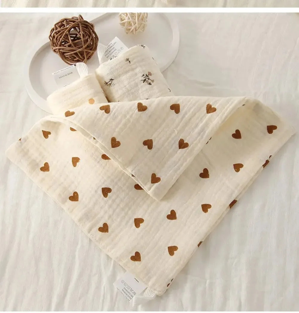 A stack of soft beige cotton gauze burp cloths with small brown heart patterns, resting on a white surface.