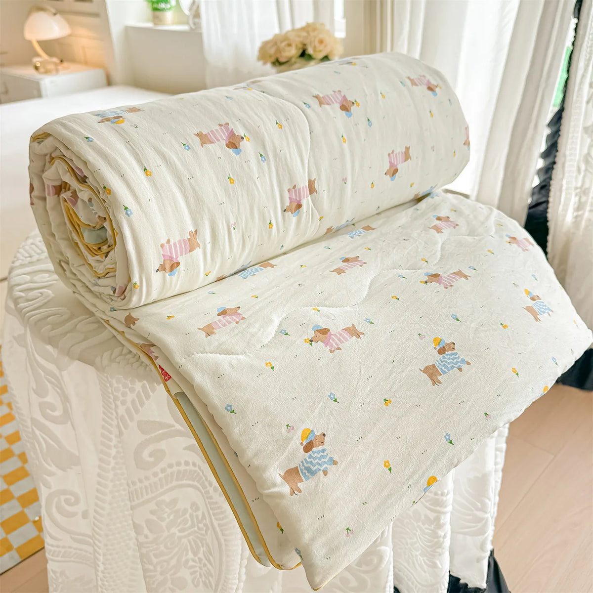 A cream-colored lightweight quilt with a pastoral print featuring small animals and flowers, rolled up on a white surface.