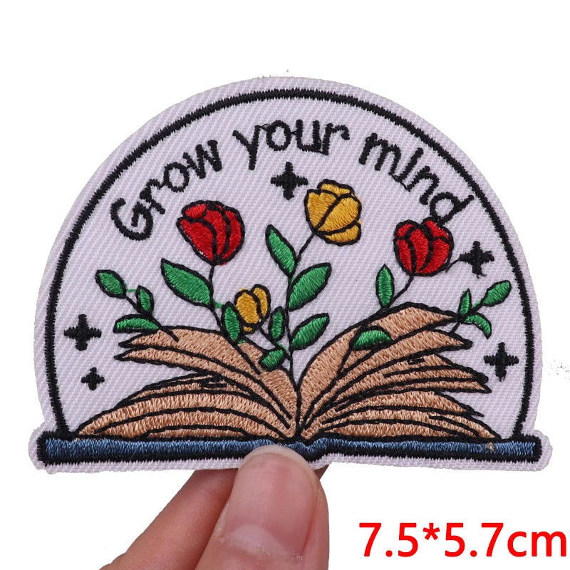 An embroidered patch featuring an open book with flowers and stars on a light pink background.