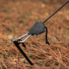 A close-up of a black tent rope tensioner with a ratchet pulley hook resting on brown, dry ground.