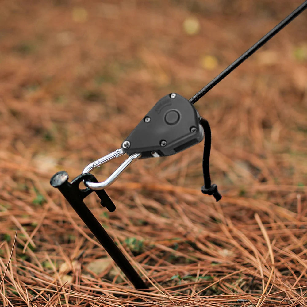 A close-up of a black tent rope tensioner with a ratchet pulley hook resting on brown, dry ground.
