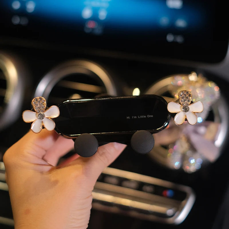 A hand holds a black gravity car phone holder with crystal flowers, positioned in a car interior.