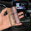 A hand holds three gold and pink refillable glass perfume atomizers in a car interior setting.