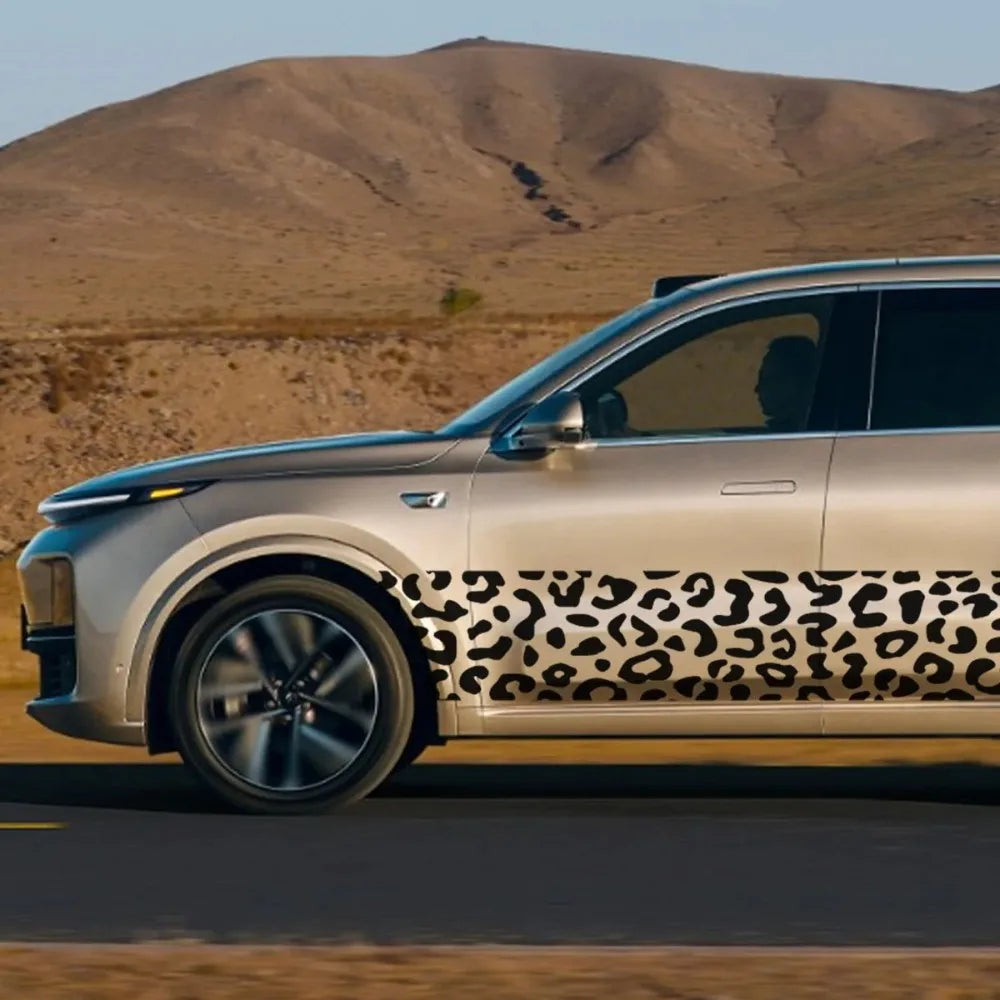 A silver car with a leopard print decal strip on the side driving on a road with dry hills in the background.