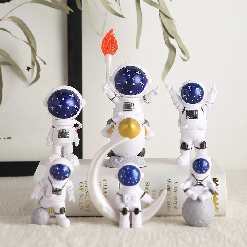 A set of handcrafted astronaut figurines in white and blue resin, including a figure with a red flame, displayed on a book.