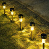 A row of modern gold-toned solar garden lantern lights illuminating dry grass on a paved walkway.