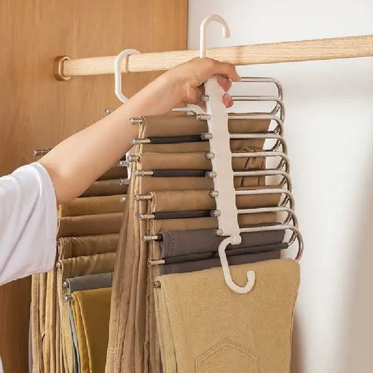 A hand is hanging a tan fabric garment onto a multi-layered stainless steel pants hanger.
