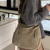 A woman holds a beige matte leather crossbody hobo bag against a window with a white cabinet and plaid skirt visible.