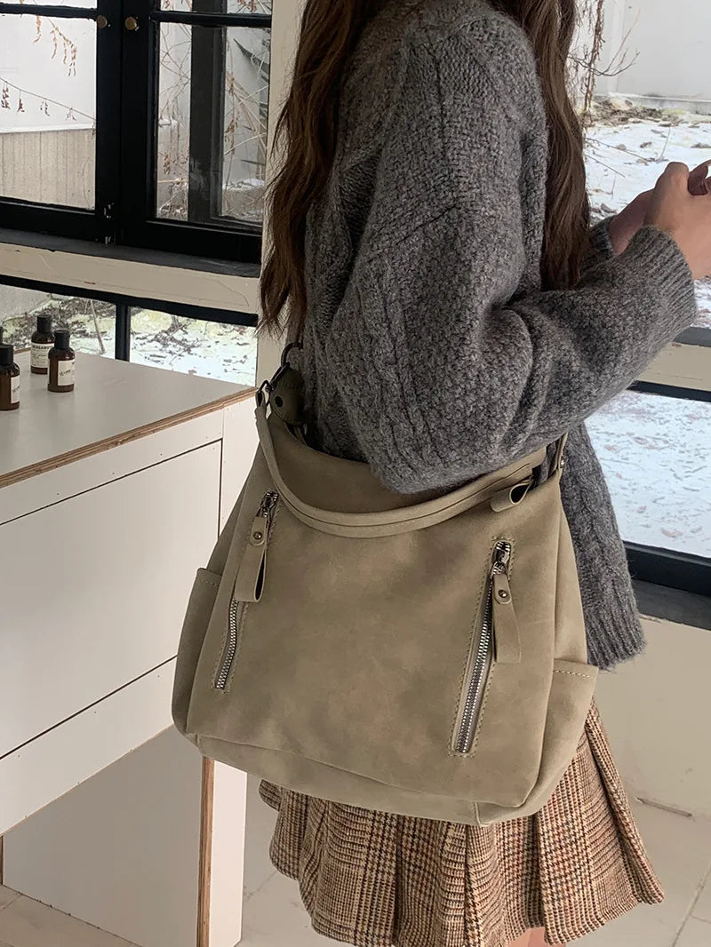 A woman holds a beige matte leather crossbody hobo bag against a window with a white cabinet and plaid skirt visible.