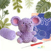 A purple crocheted elephant toy next to yarn and crochet tools with green foliage.