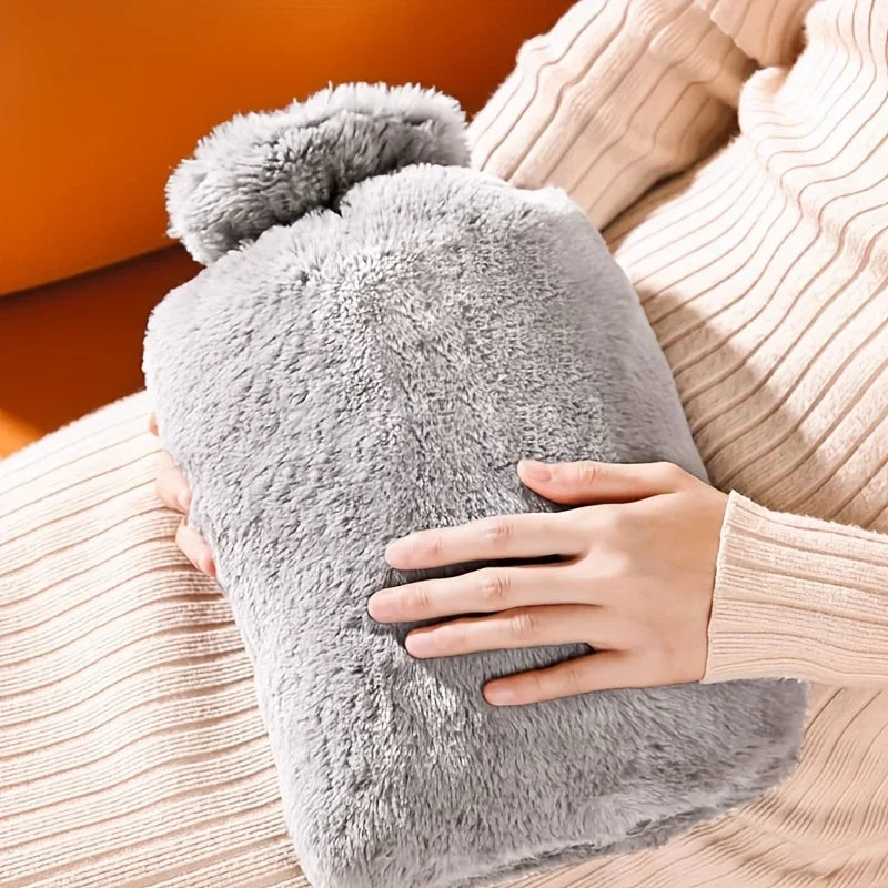 Eine Hand hält eine große graue Plüsch-Heißwasserflasche an einem beige gestrickten Stoff.