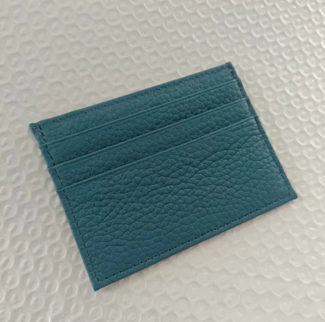 Porta tarjetas de cuero genuino y delgado color verde azulado con acabado texturizado sobre una superficie blanca.