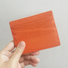 A hand holds an orange genuine leather slim card holder wallet with a textured surface.