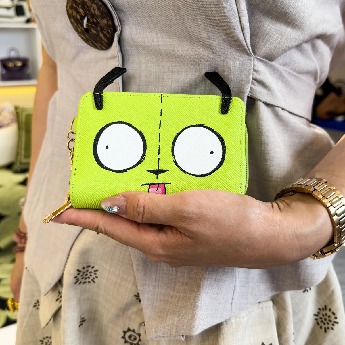 A hand holds a bright lime green cartoon print compact wallet with black keychain charms.