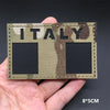 A hand holds a rectangular tactical patch featuring the word ITALY in camouflage print on a fabric background.