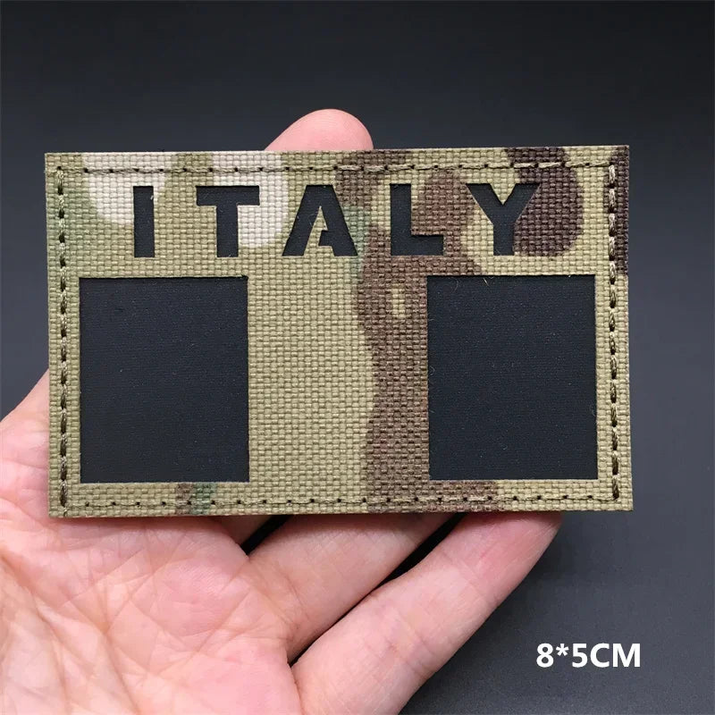A hand holds a rectangular tactical patch featuring the word ITALY in camouflage print on a fabric background.