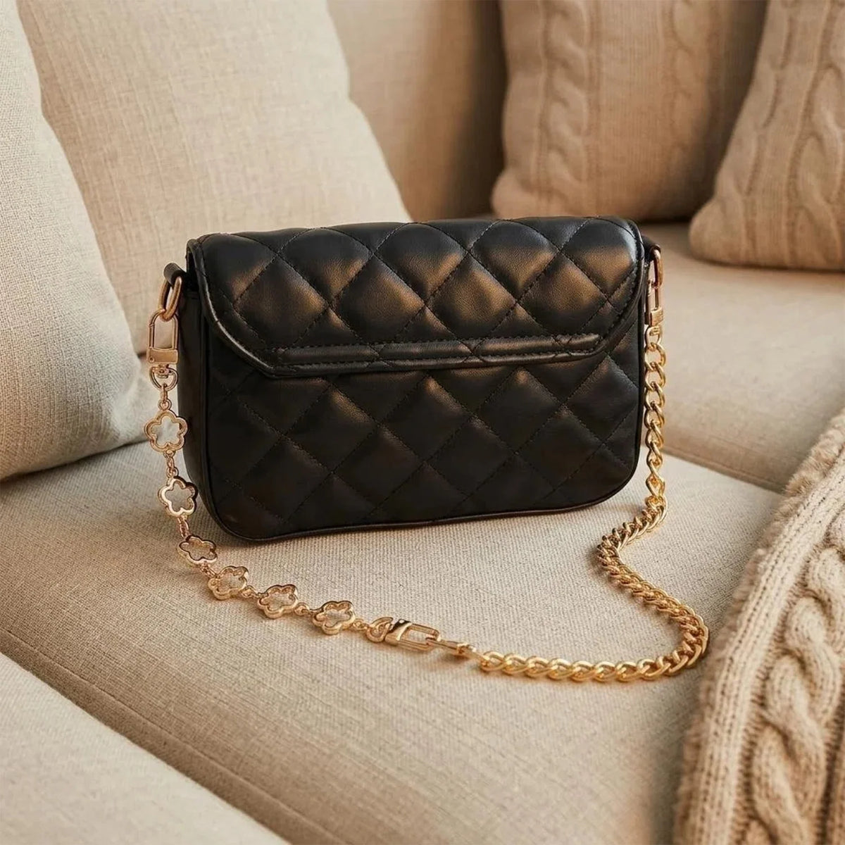 A black quilted flap bag with a gold chain extender rests on a beige sofa cushion.