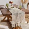 A beige tasseled table runner is laid across a wooden table set with gold goblets and fruit.