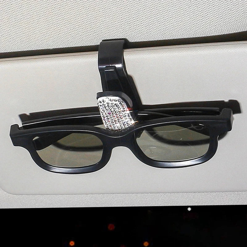 Black sunglasses with a rhinestone clip resting on a light-colored car visor.