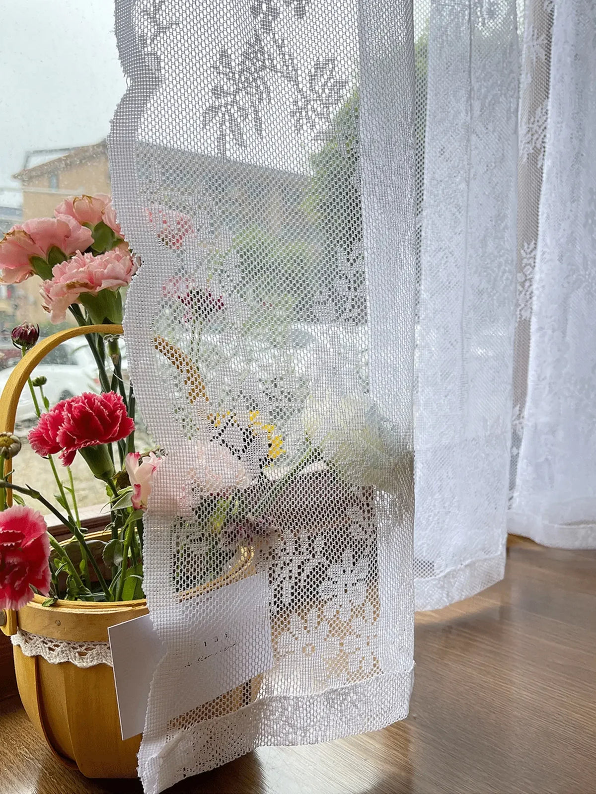 Un rideau blanc transparent avec broderie florale pend à côté d'un arrangement de fleurs roses et rouges en pot.