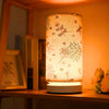 A Nordic retro USB table lamp with a sheer fabric shade featuring nature-themed patterns, illuminated on a wooden shelf.