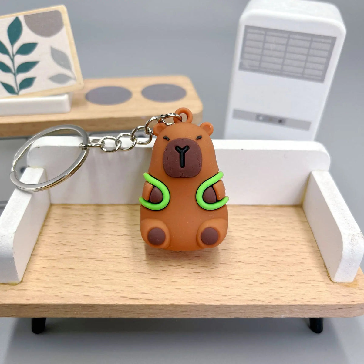 An adorable brown capybara keychain charm hanging from a silver chain, displayed on a wooden tray.