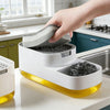 A hand is placing a gray sponge into the dispenser of a white countertop soap dispenser with a yellow base.
