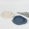 A set of two smooth silicone coasters, one beige and one blue-gray, are displayed next to a sprig of eucalyptus.