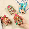 A hand holds a DIY book nook fridge magnet kit featuring an illuminated miniature house and surrounding natural elements.