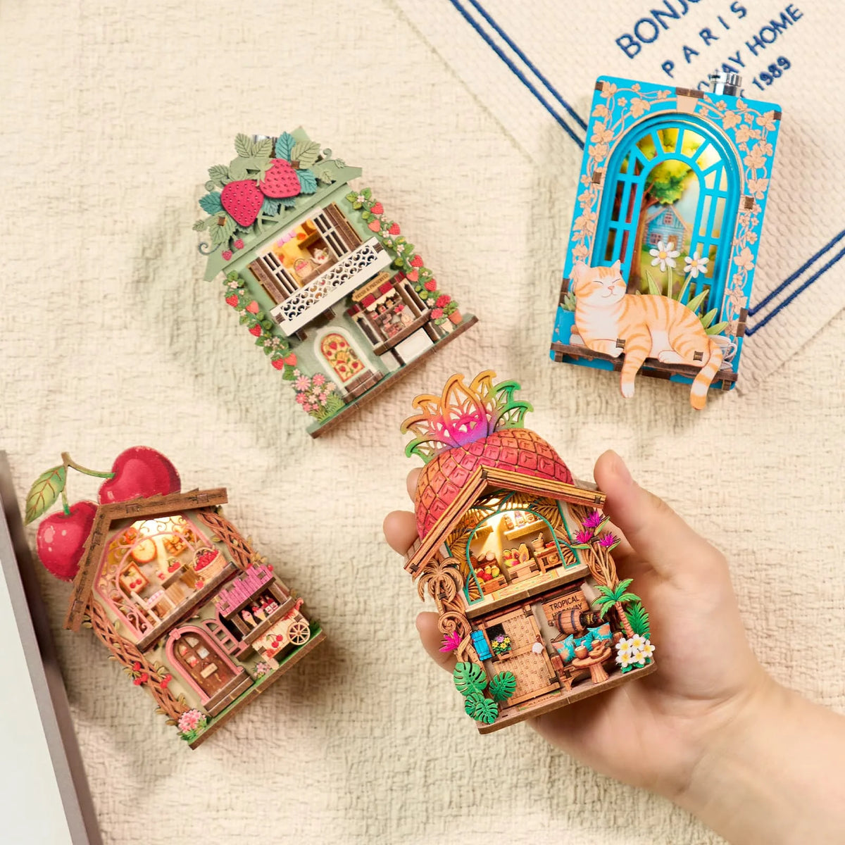 A hand holds a DIY book nook fridge magnet kit featuring an illuminated miniature house and surrounding natural elements.