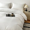 A white brushed duvet cover with a folded white duvet and a bedside lamp on a wooden surface.
