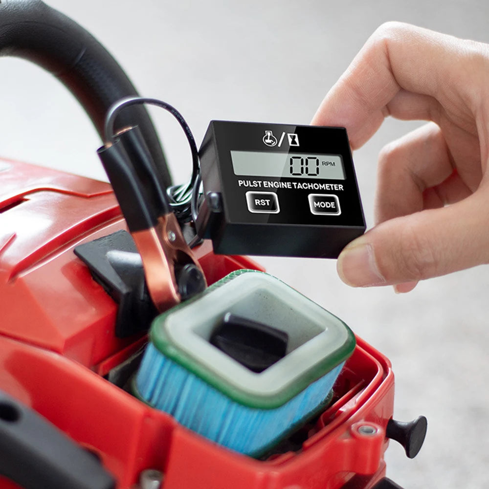 A hand holds a digital tachometer and hour meter device attached to a red power tool.